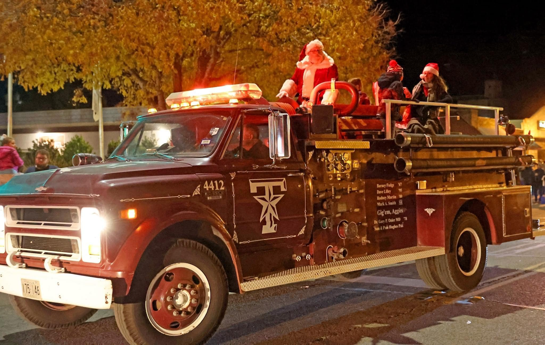 Santa and helpers on a vintage maroon fire truck with lights, in a nighttime parade.