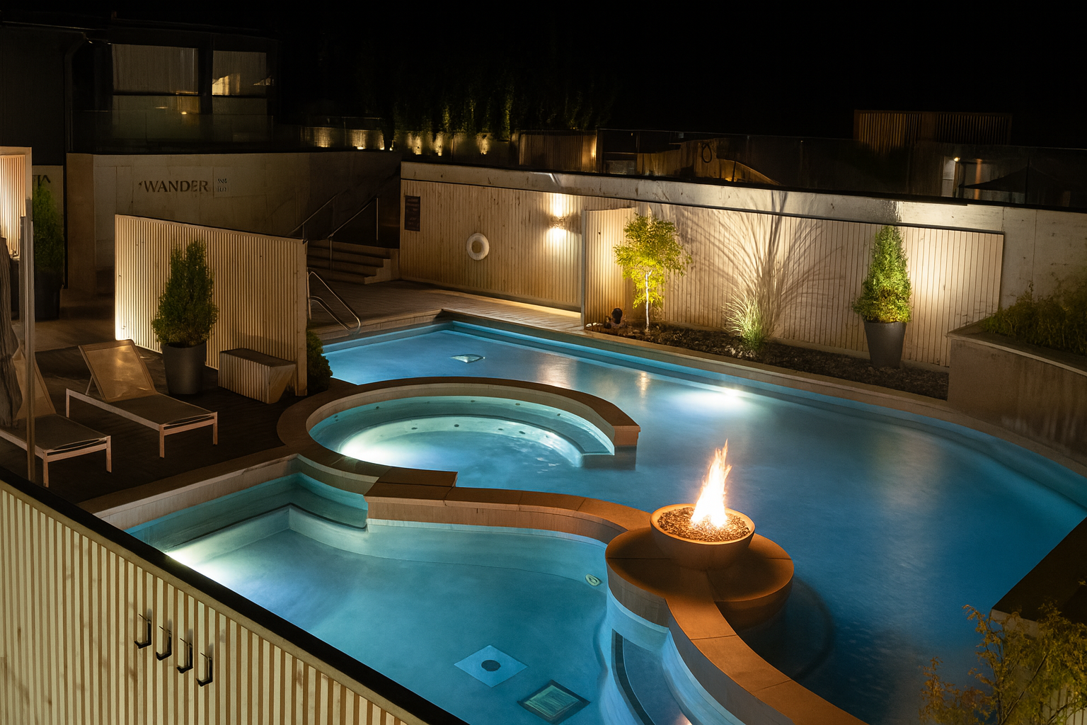 Rooftop pool and jacuzzi at night, lit by underwater lights, fire pit, and wall lighting.