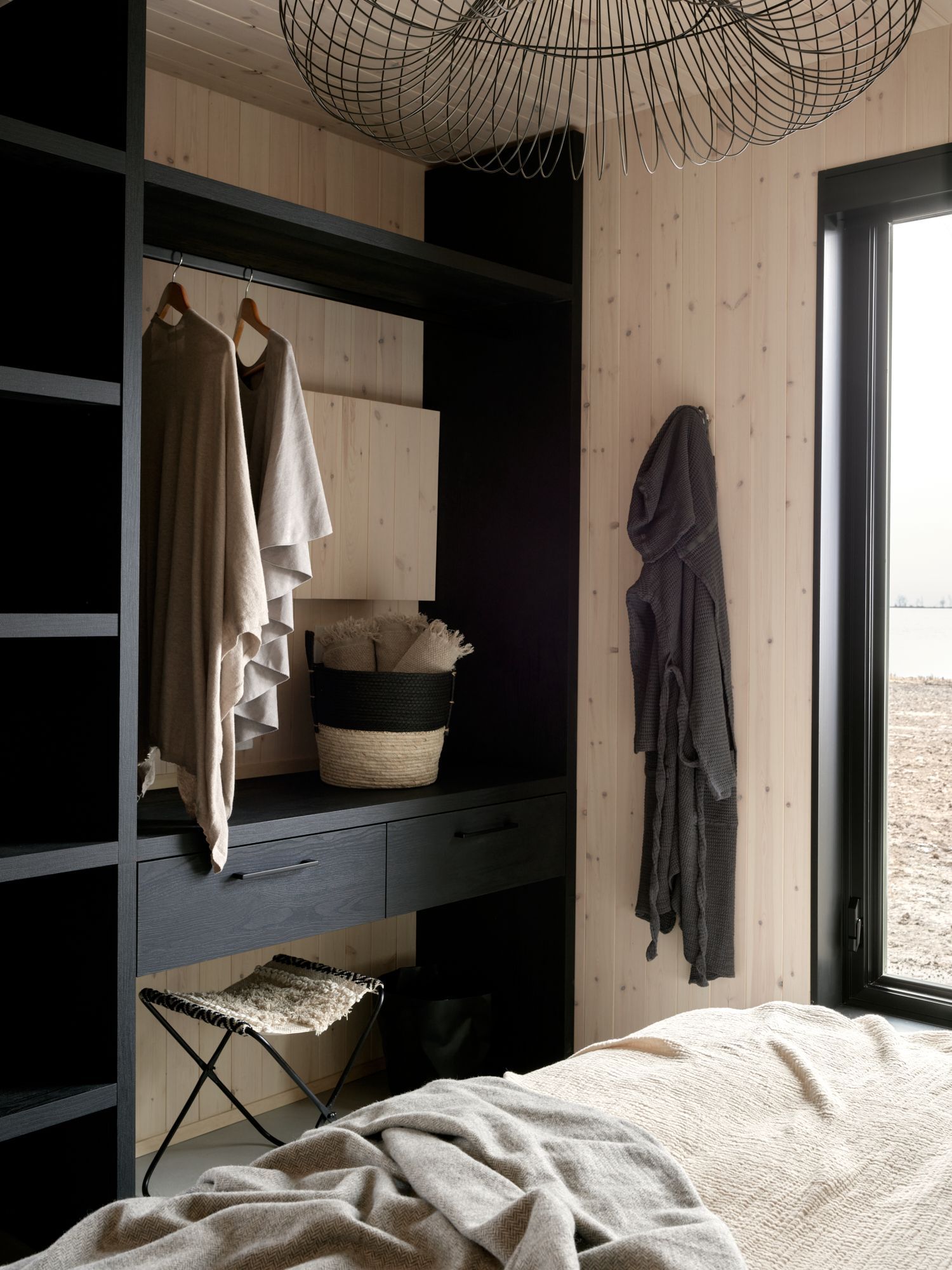 Closet with dark shelving, hanging clothes, robe, and a small stool in a bedroom with wood-paneled walls.