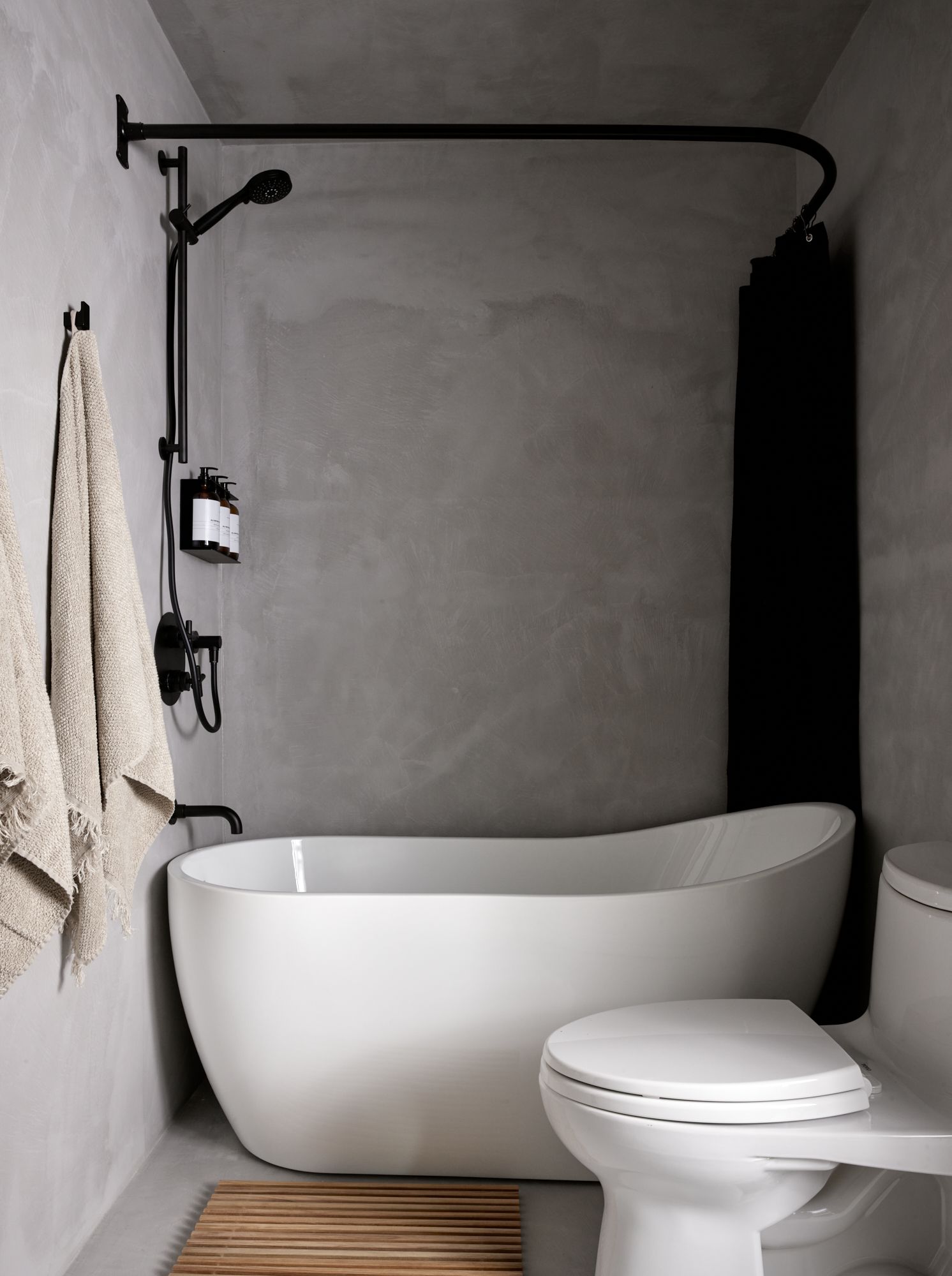 Modern gray bathroom with white tub, black shower fixtures, and toilet. Beige towels on a hook.