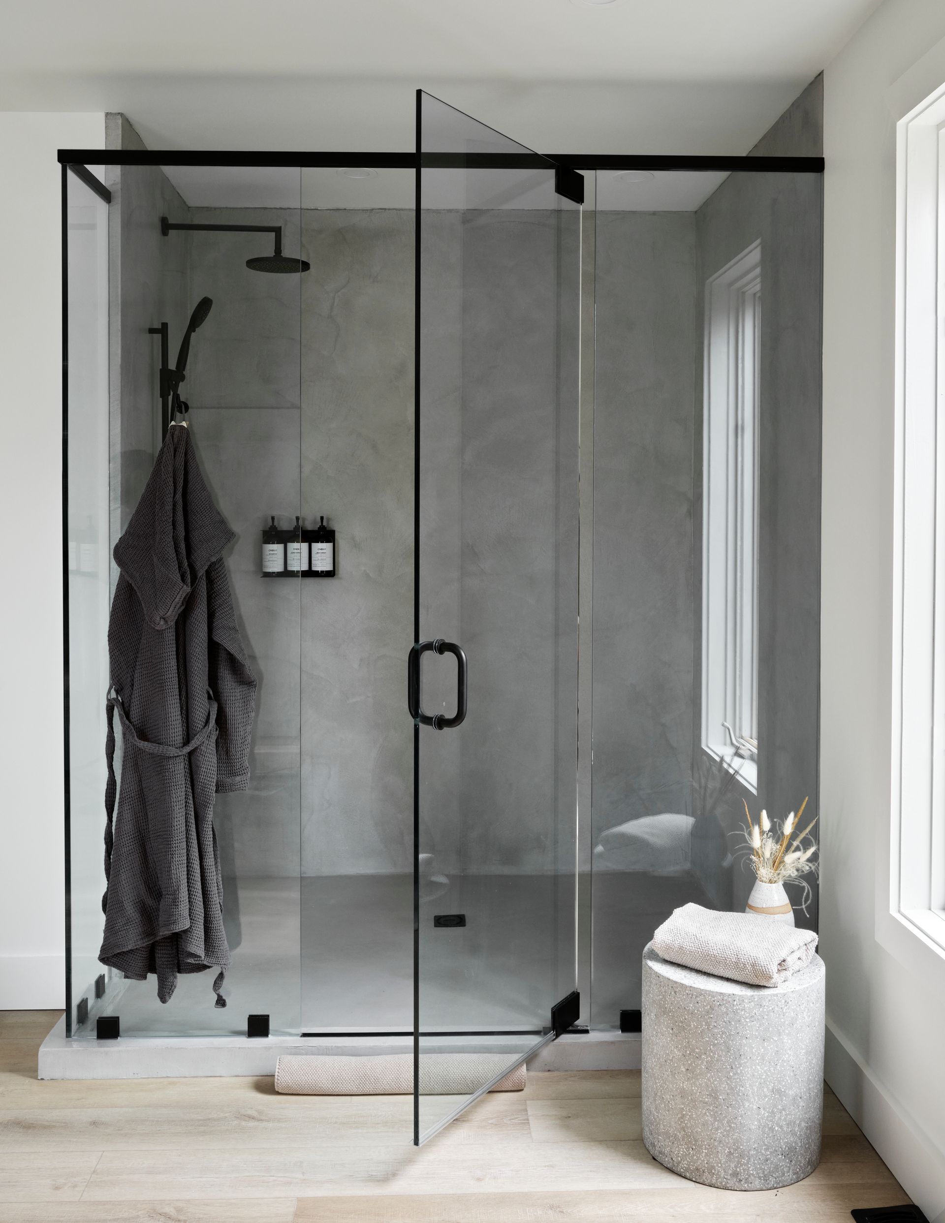 Modern walk-in shower with gray walls and black-framed glass doors, a robe hangs, towels on a stone stool.