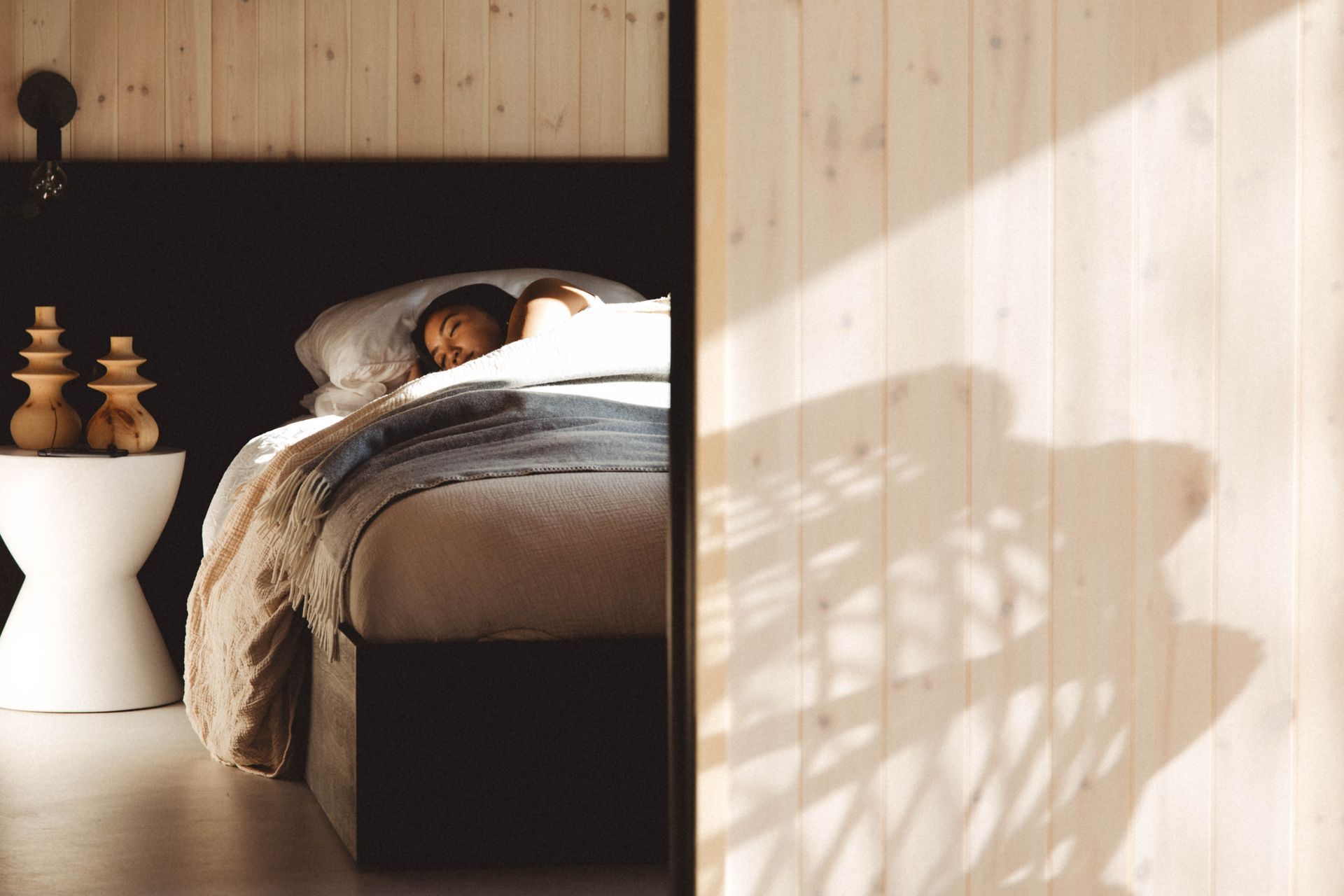 Person asleep in bed in a wooden-paneled room. Sunlight streams through a door.