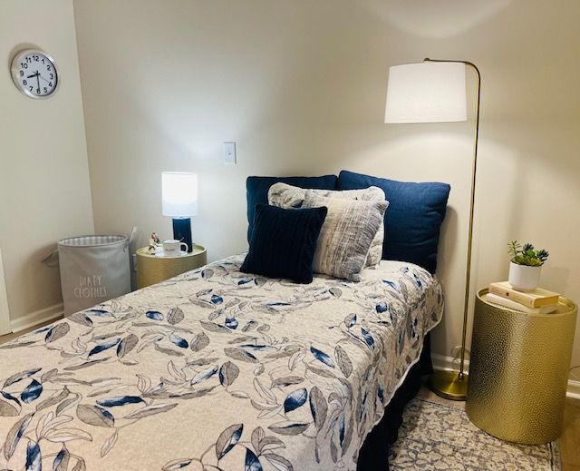 A bedroom with a bed , lamp , nightstand and a clock on the wall.