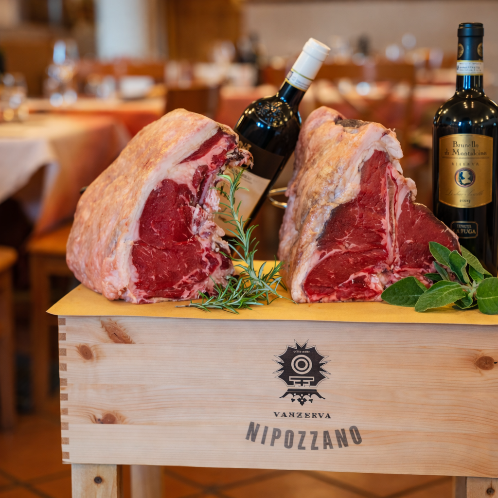 Due grandi bistecche di carne rossa, appoggiate su una scatola di legno con due bottiglie di vino, rosmarino e salvia.