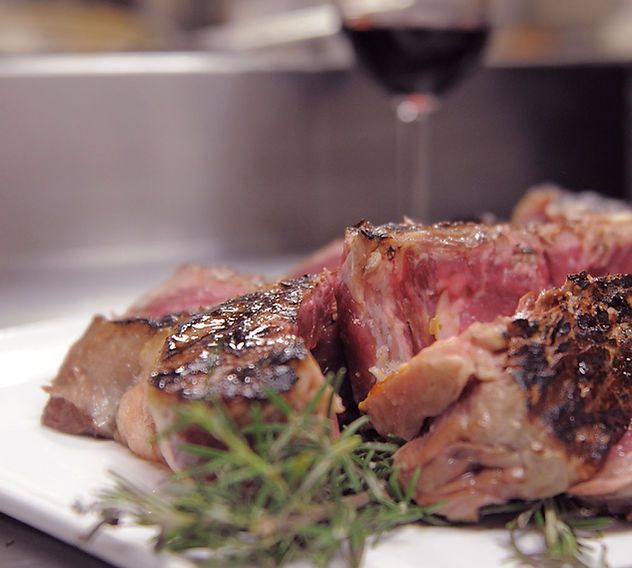 Bistecca tagliata alla griglia su un piatto bianco con rosmarino e un bicchiere di vino rosso sullo sfondo.
