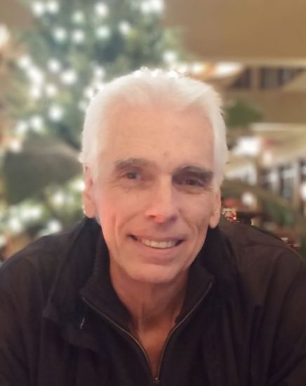 A smiling person with light-colored hair wears a dark zip-up jacket in front of a blurry, illuminated holiday tree.