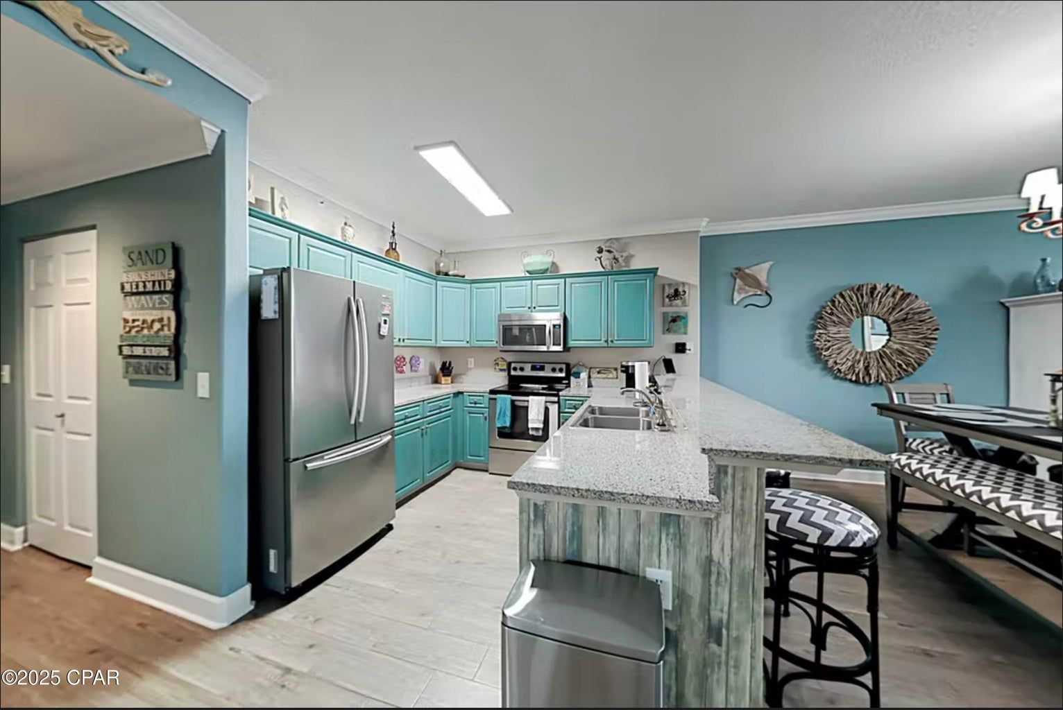 A bright kitchen with teal cabinets, stainless steel appliances, a granite-topped island, and a dining area with a mirror.