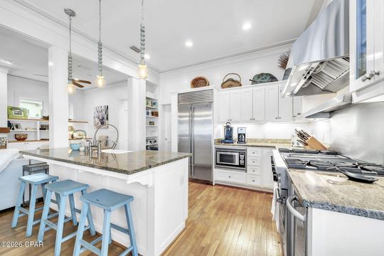 A white kitchen featuring a granite-topped island with three light blue stools, stainless steel appliances, and wood floors.