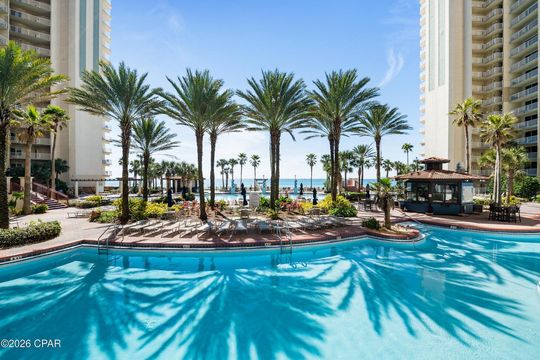 A bright, sunny swimming pool courtyard framed by tall resort buildings with palm trees and a view of the ocean.
