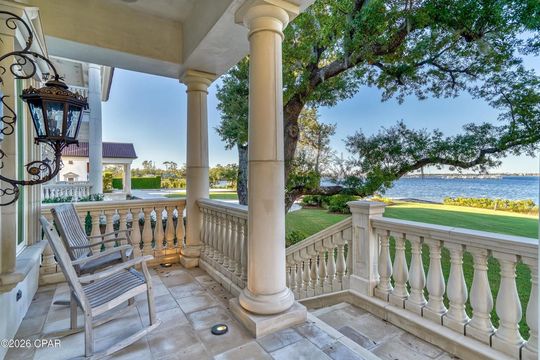 A stone porch with two rocking chairs overlooking a grassy lawn, large trees, and a body of water under a clear blue sky.