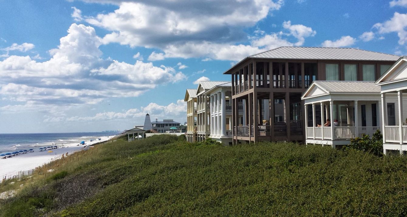 A row of beachfront houses with porches on a grassy dune overlooks a sandy beach under a cloudy blue sky.