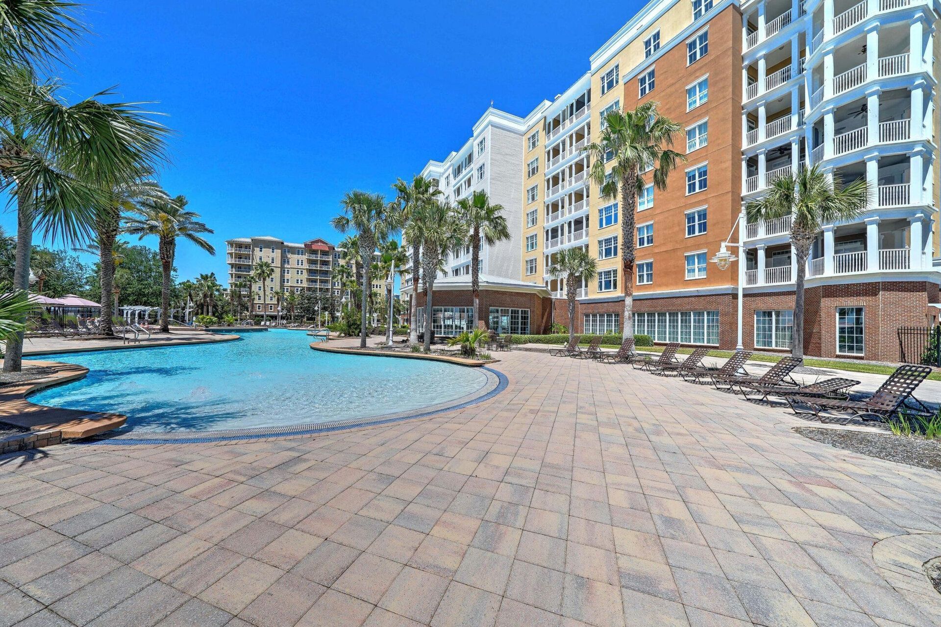 A sunny pool deck paved with stone pavers leading to a large resort building and palm trees under a clear blue sky.