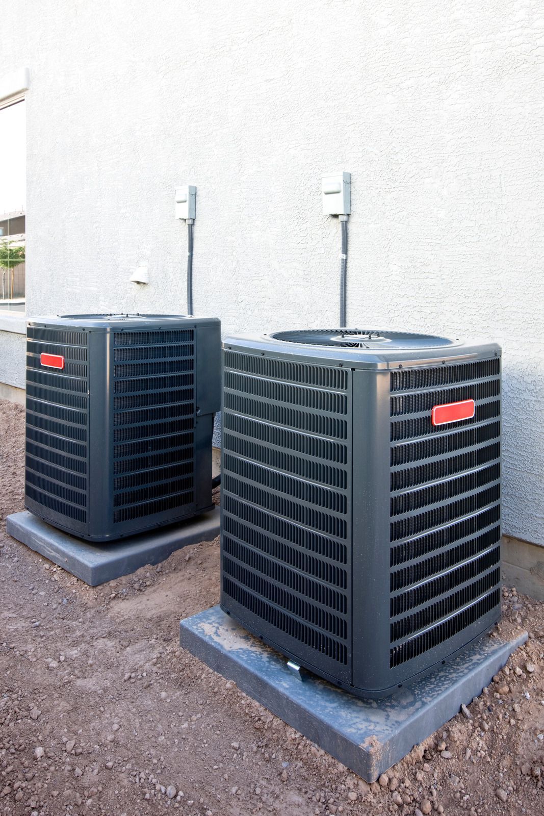 Two outdoor air conditioning units on concrete pads near a white wall.