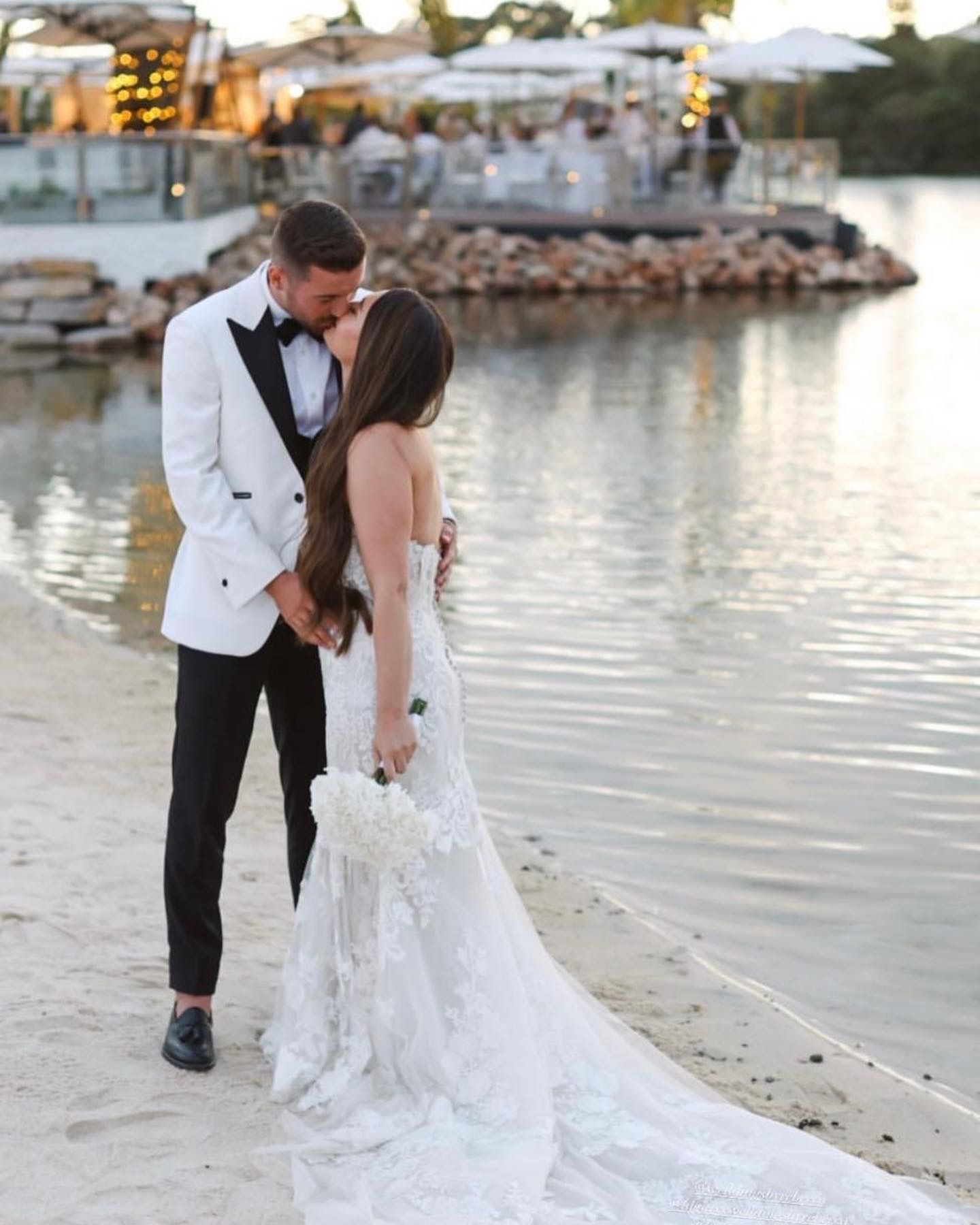 A bride and groom are kissing on the beach near the water.