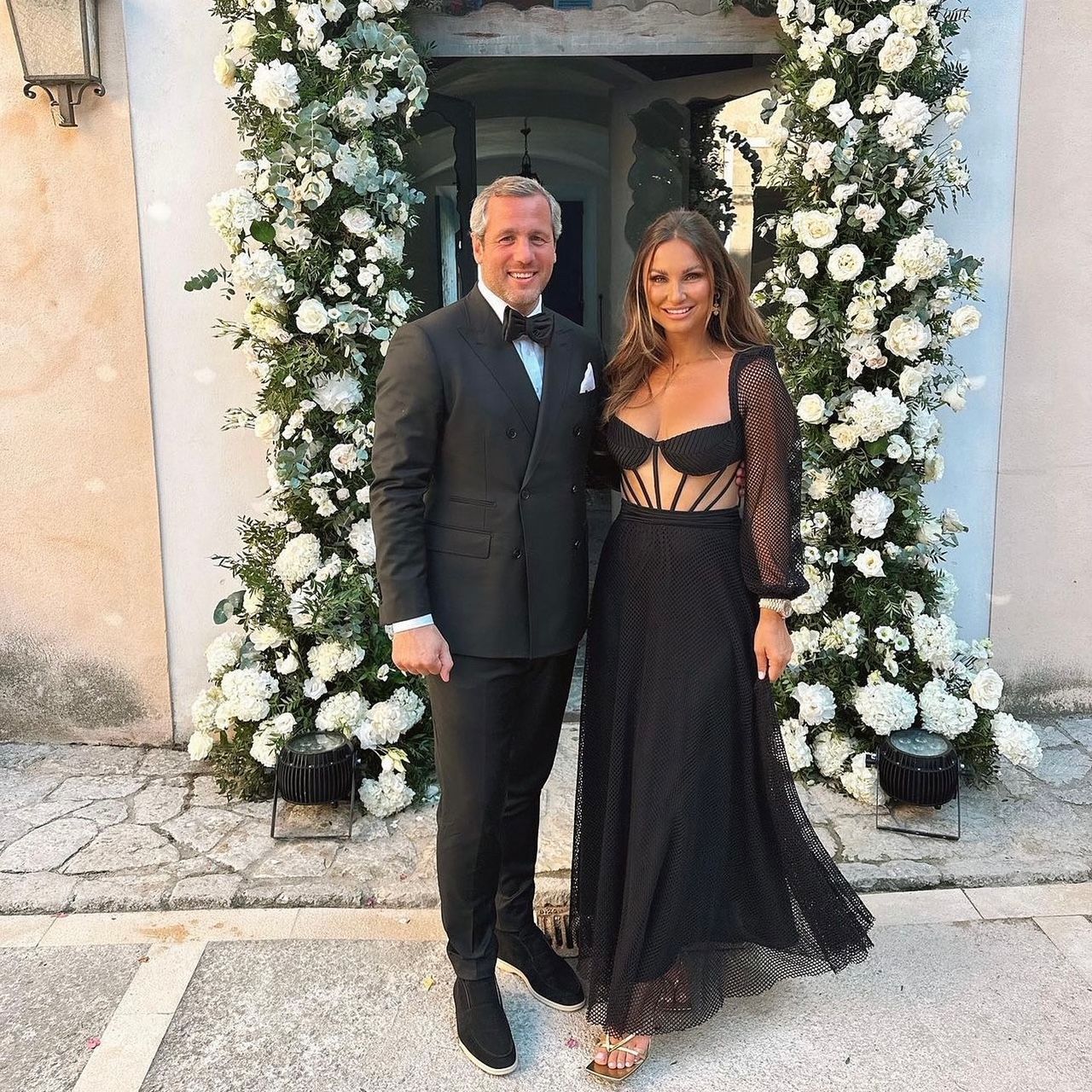 A man in a suit and a woman in a black dress are posing for a picture in front of flowers.