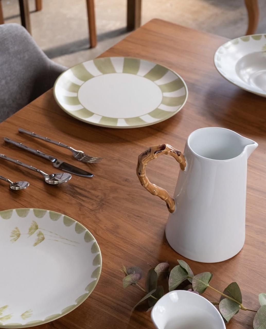 Juego de mesa de madera con platos, cubiertos, jarra blanca y vegetación.