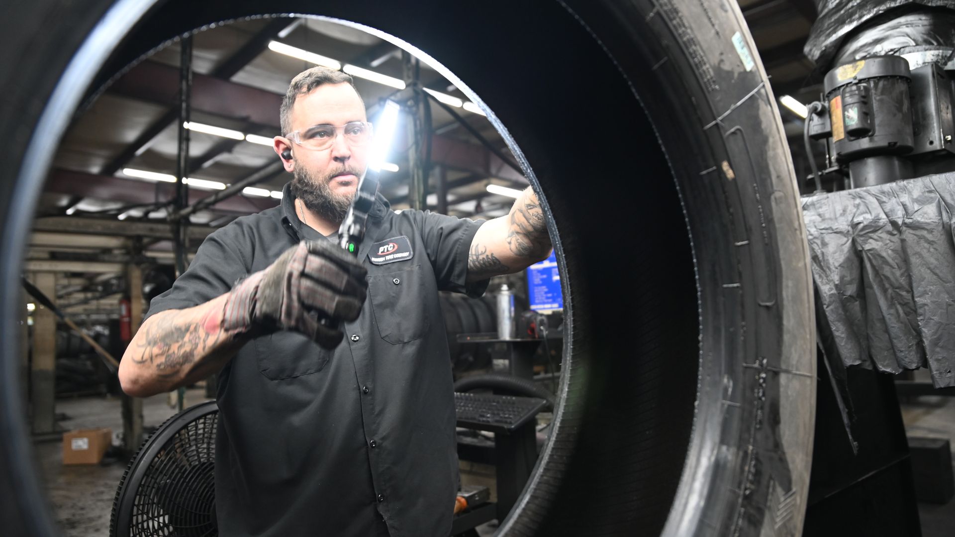 Man inspecting a large tire inside a workshop, wearing gloves, holding a tool.