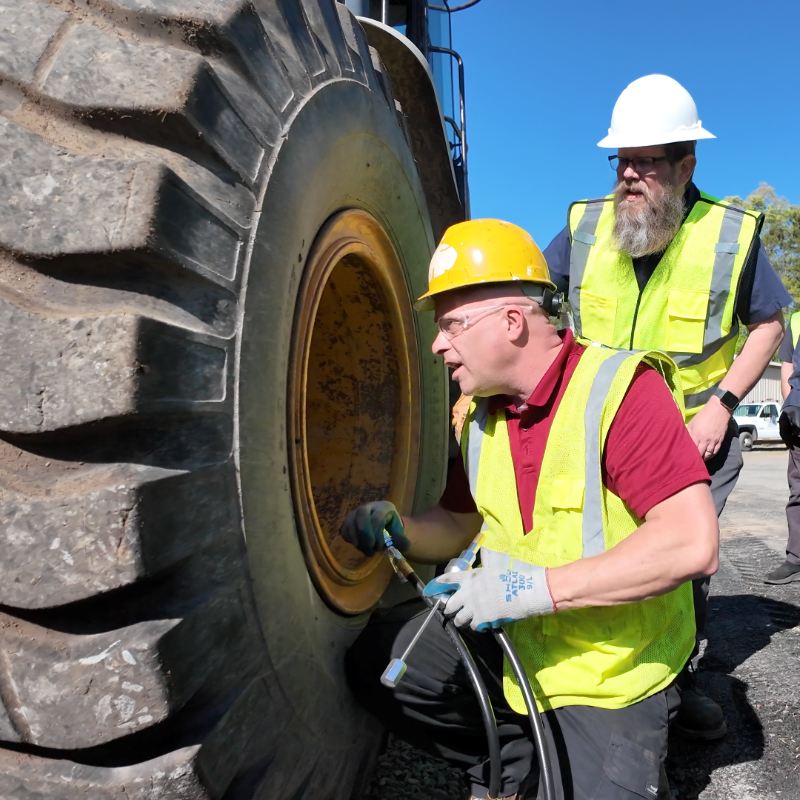Two people inspect a large tire on a construction vehicle. One kneels, using a tool; both wear safety vests and helmets.