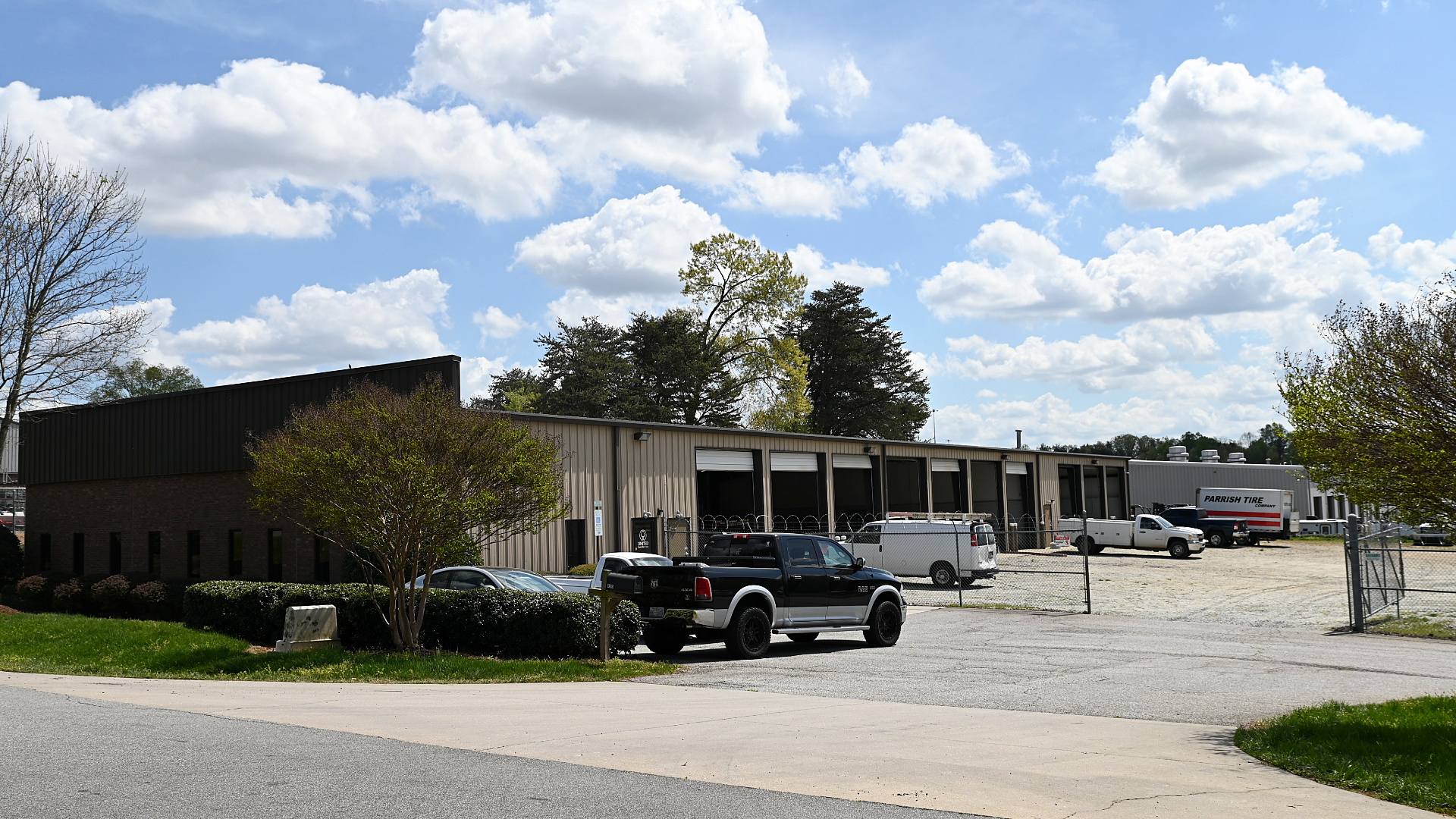 A long, low industrial warehouse with loading bays, a gravel lot, and parked vehicles under a partly cloudy sky.