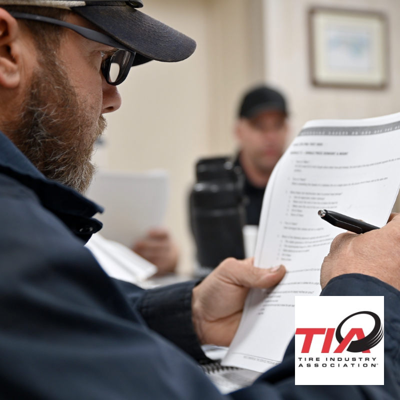 Man in cap and glasses examines paperwork, another person in background. Tire Industry Association logo in corner.