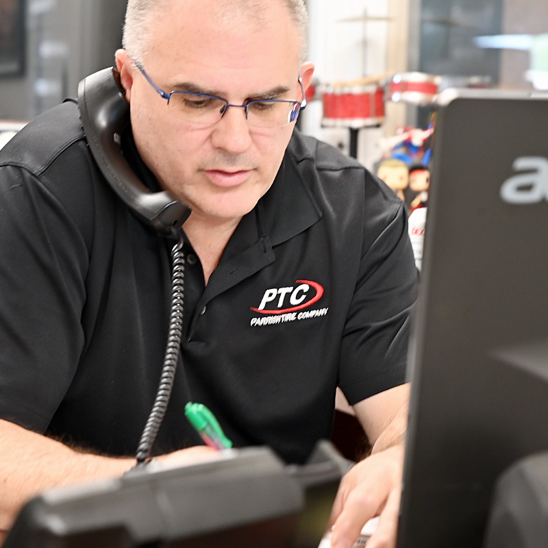 Man wearing glasses on the phone at a desk, looking at a computer; PTC logo on his black shirt.
