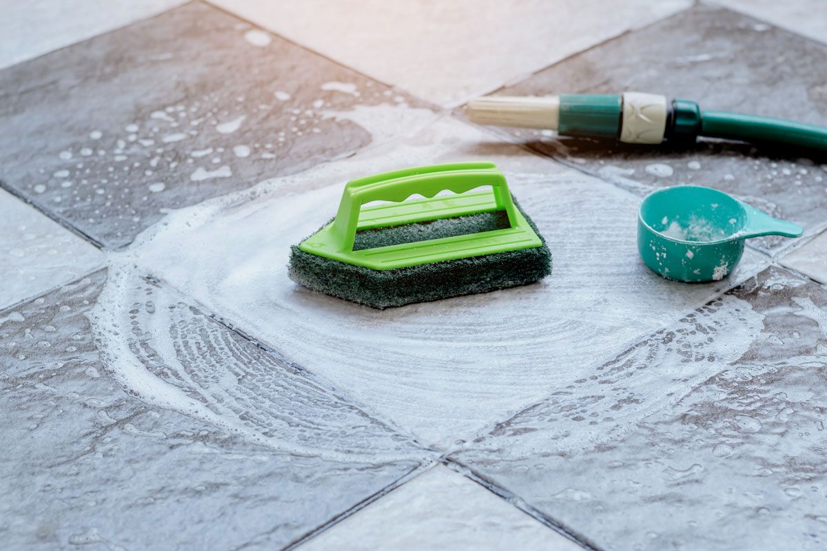 A sponge , brush , and measuring cup are sitting on a tiled floor.
