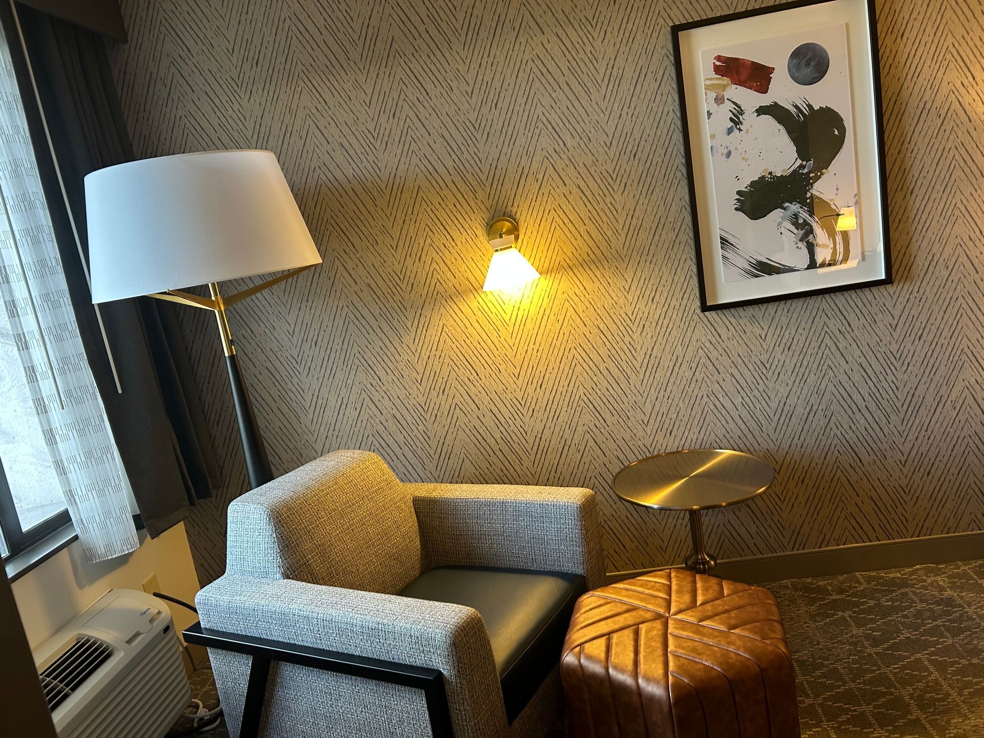 A hotel room with a chair , lamp , table and ottoman.