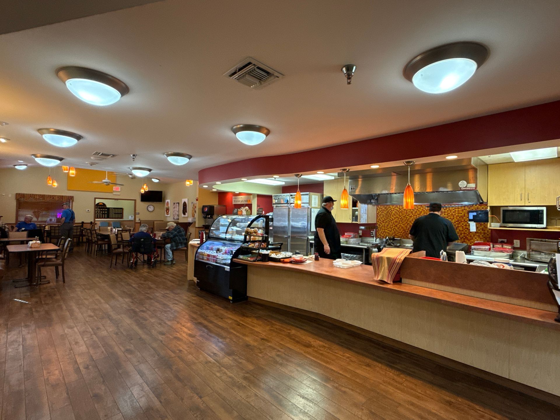 Restaurant interior: diners seated, kitchen staff preparing food, display case with food items.