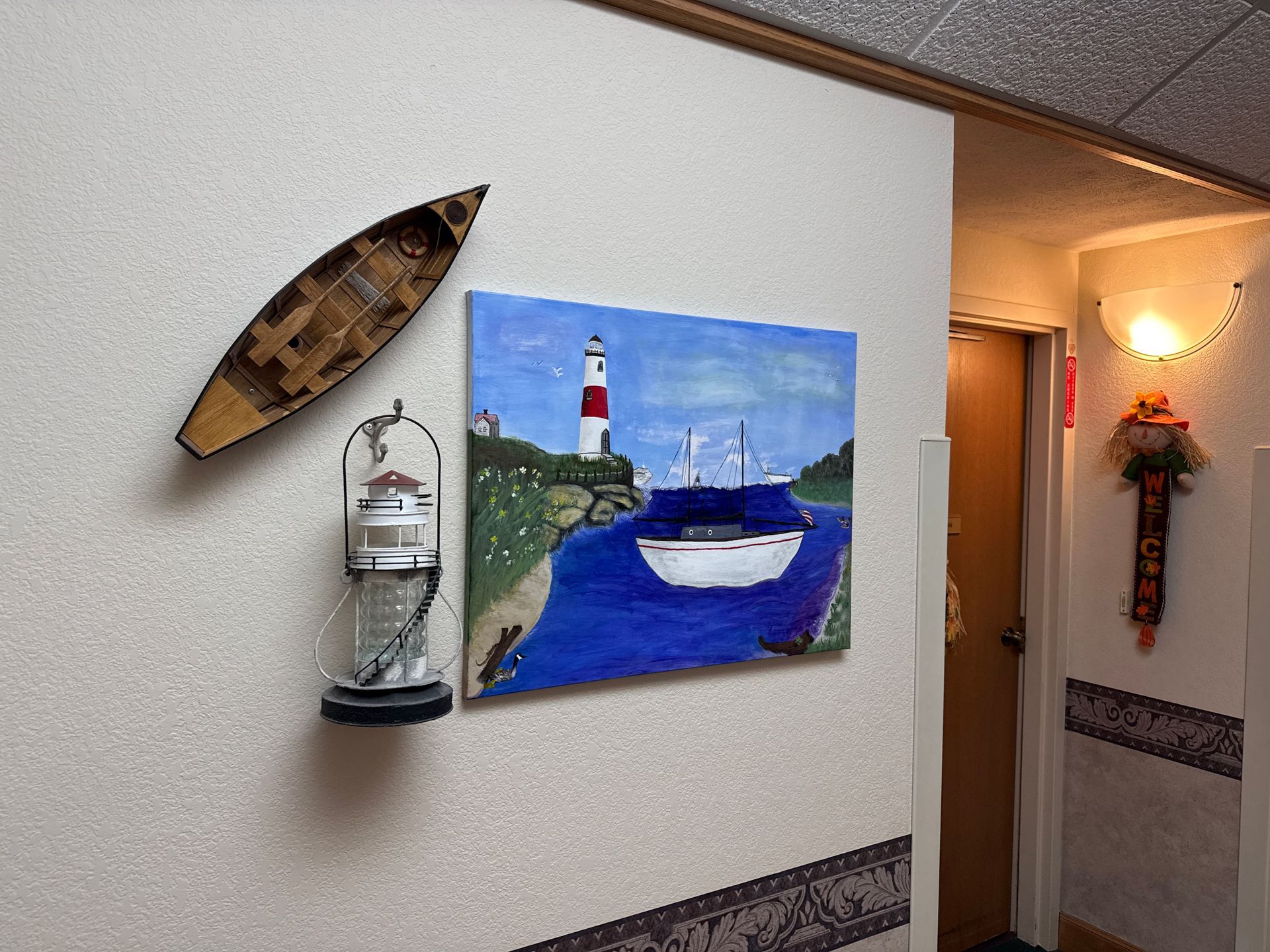 Coastal-themed wall decor: a painting of a boat and lighthouse, a wooden boat, and a lantern on a textured wall.