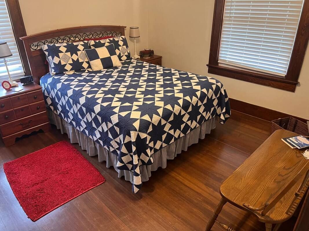 Bedroom with wooden floors, bed with blue and white star quilt, red rug, and small wooden table.
