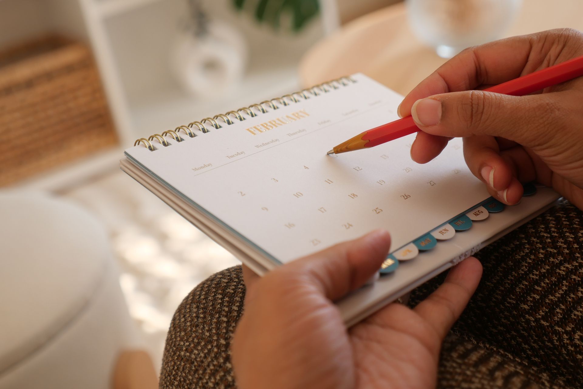 Person using a red pencil to write in a spiral-bound calendar, presumably scheduling tasks.