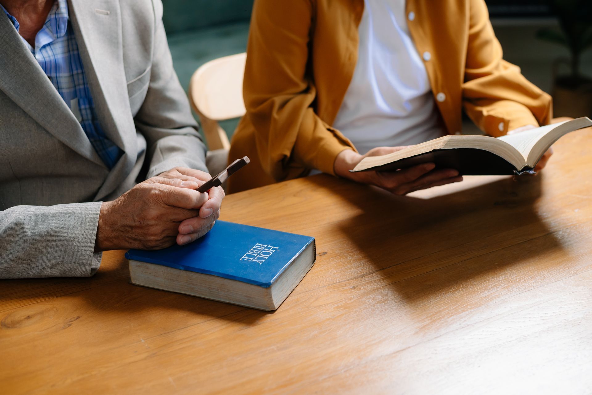 Two people at a table, one holding a book, the other with a pen and Bible.