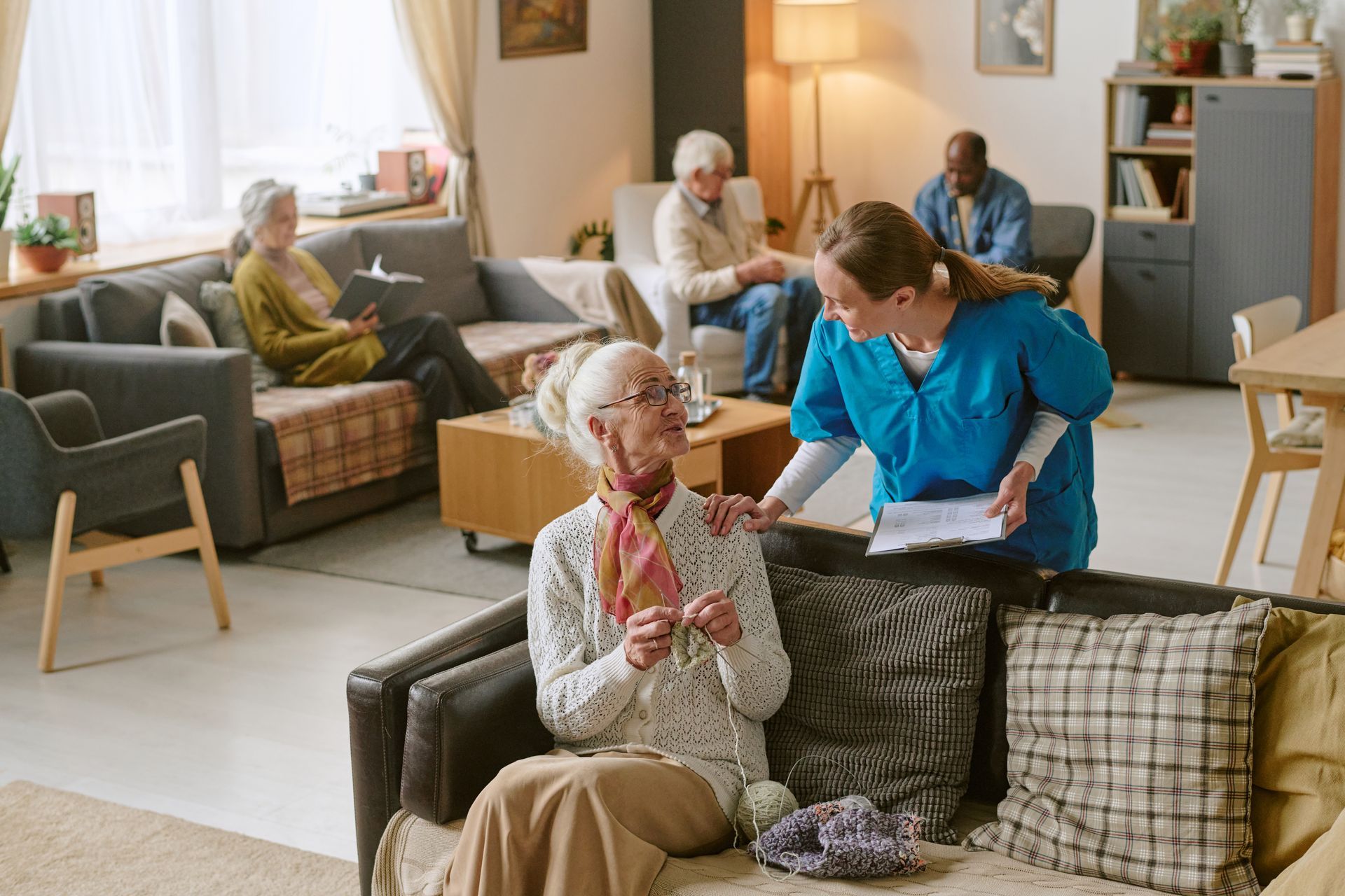 Assisted living resident knits while nurse reviews a document. Other residents sit in the living area.