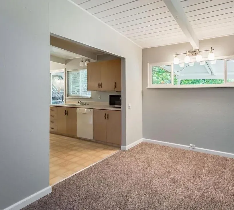 An empty room with a carpeted floor and a kitchen in the background.