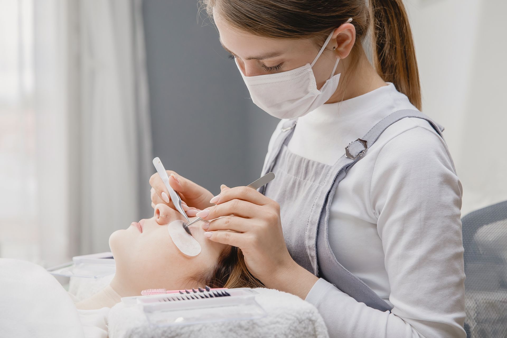 A woman wearing a mask is applying eyelash extensions to a woman 's eye.