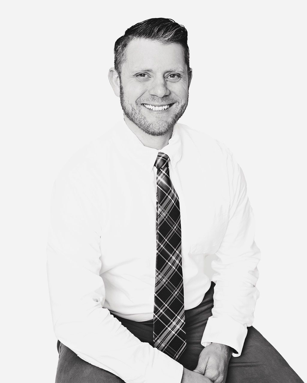 A man in a white shirt and plaid tie is sitting on a stool.