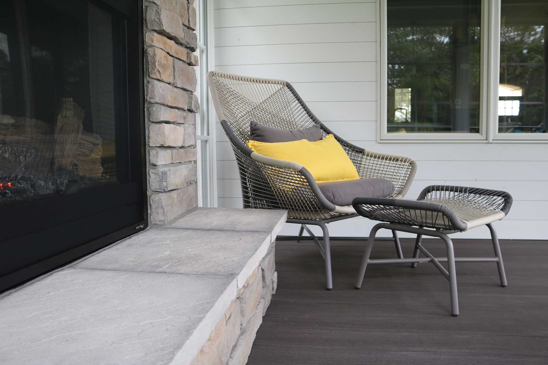 A chair and ottoman are on a porch next to a fireplace.