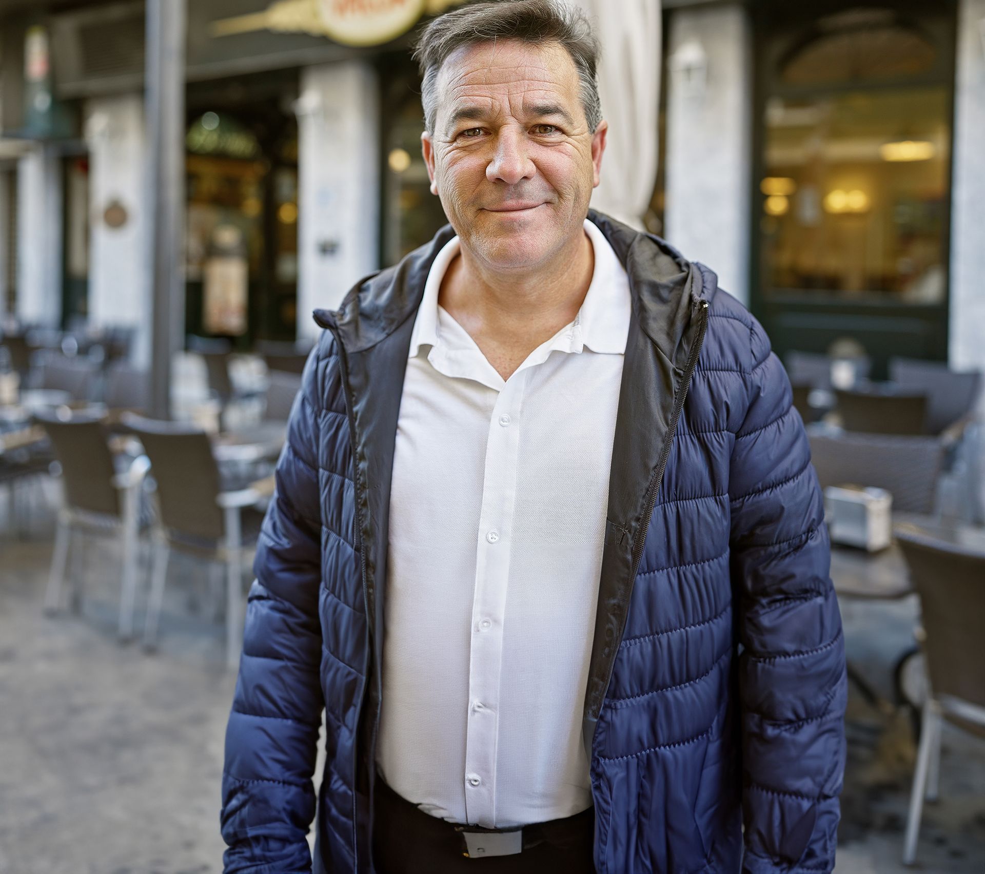 Man in blue jacket smiles outdoors in front of a cafe with tables and chairs.