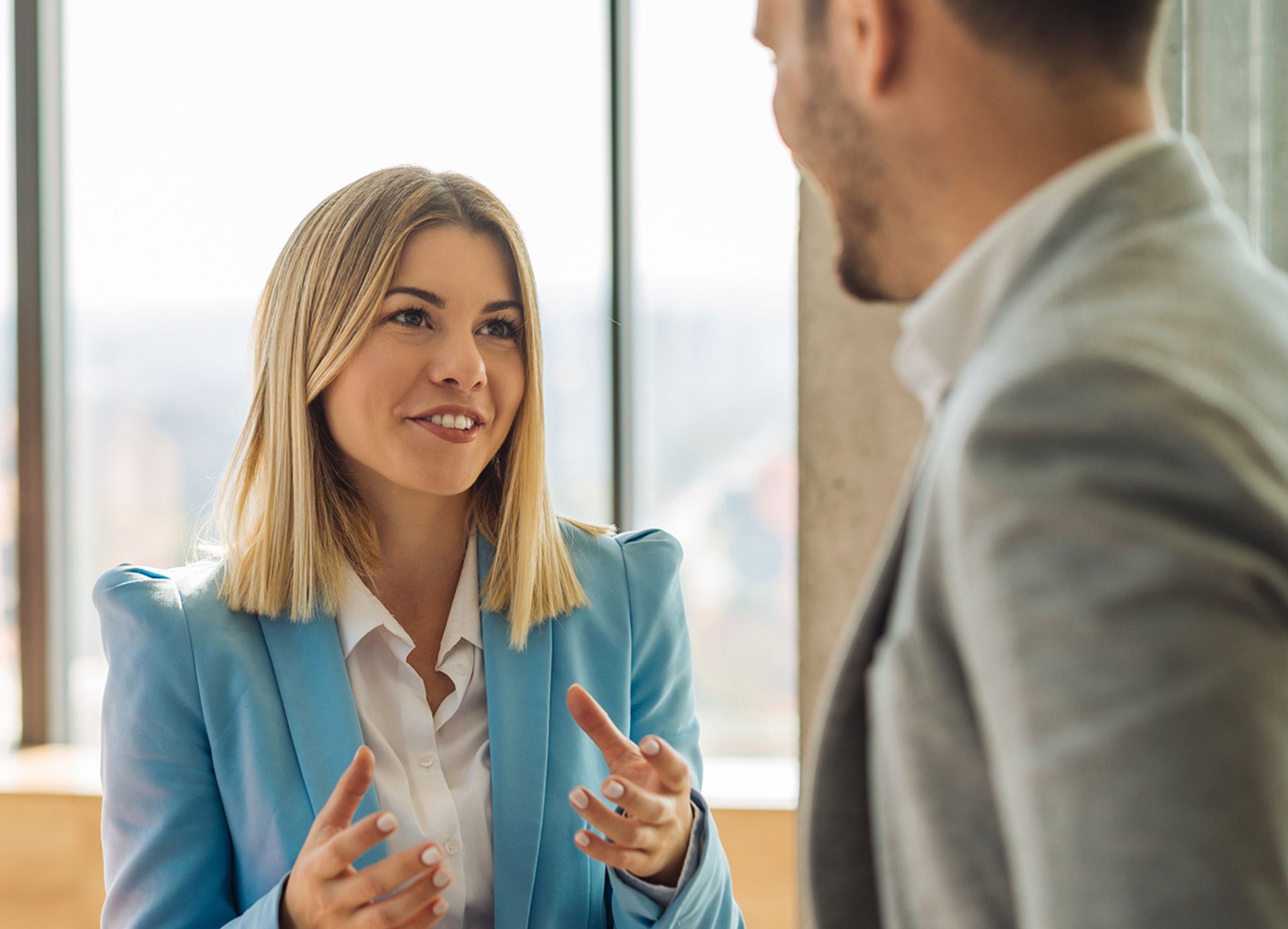 Woman in blue blazer gestures while talking to a man in a gray suit near a window.