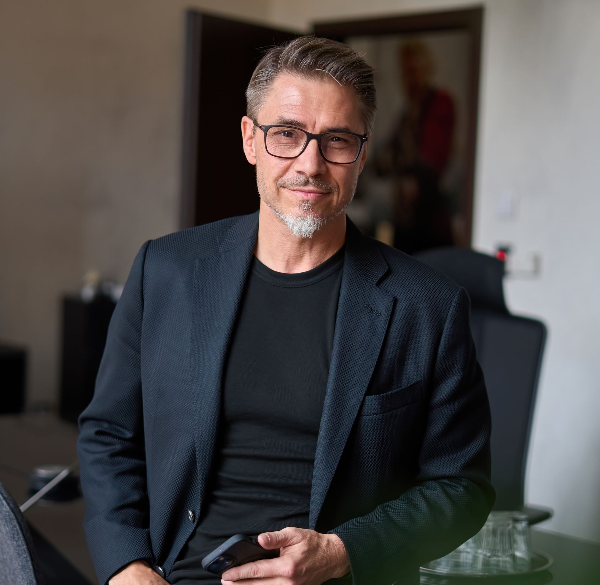 Man in glasses and blazer holding a phone, looking at the camera. Neutral expression, indoor setting.