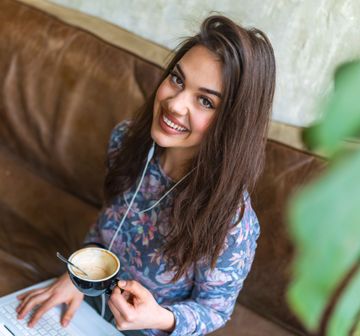 Woman smiles at the camera, holding a coffee cup and wearing earbuds while sitting on a leather couch.