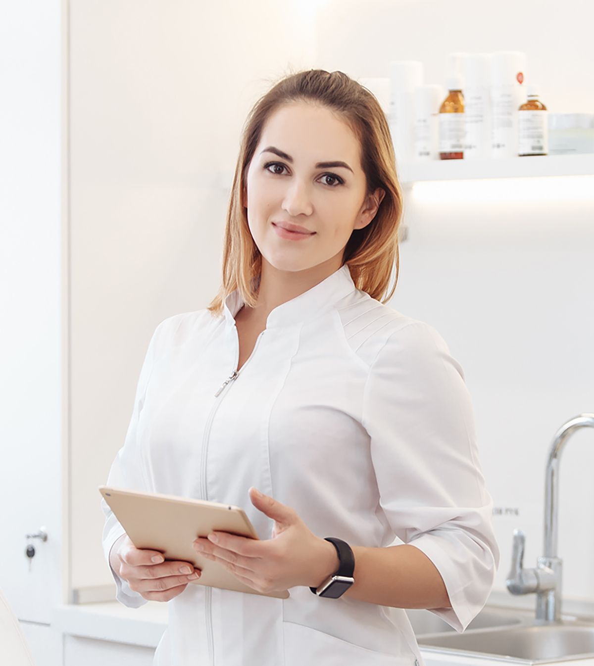 Woman in white coat holding a tablet, smiling, in a bright medical setting.