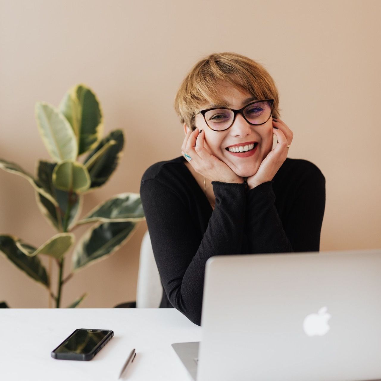 Woman wearing glasses smiles at a laptop, leaning on her hands in a home office with a plant.