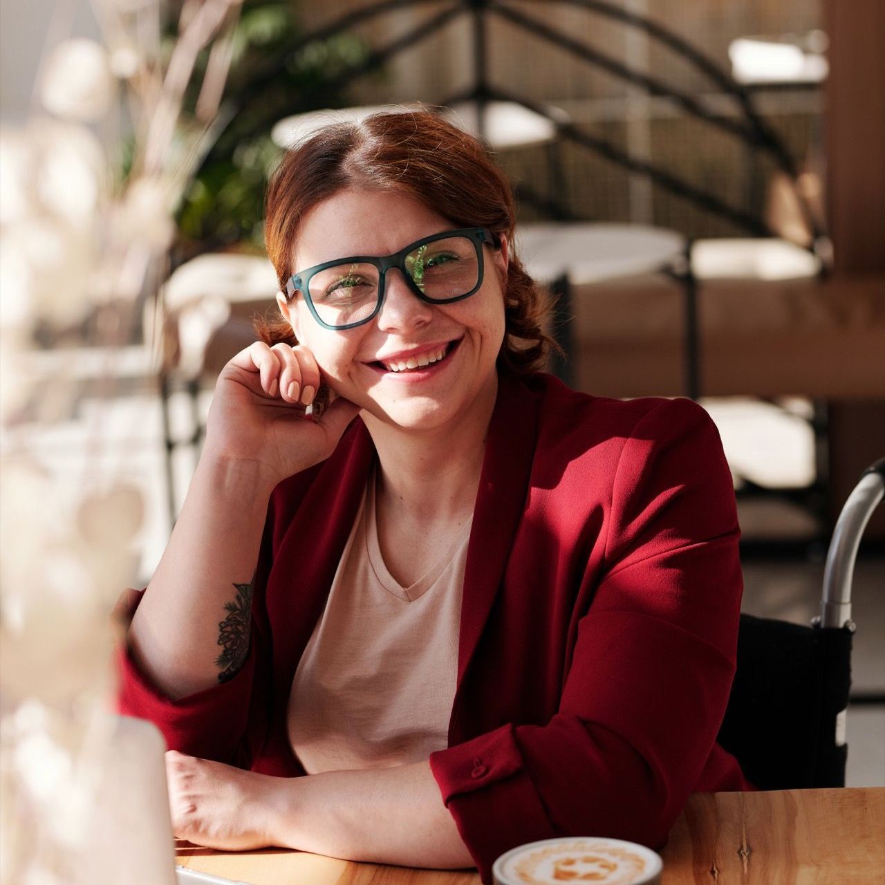 Woman with glasses, red blazer, and smile sits in a wheelchair at a table in a cafe, looking at the camera.