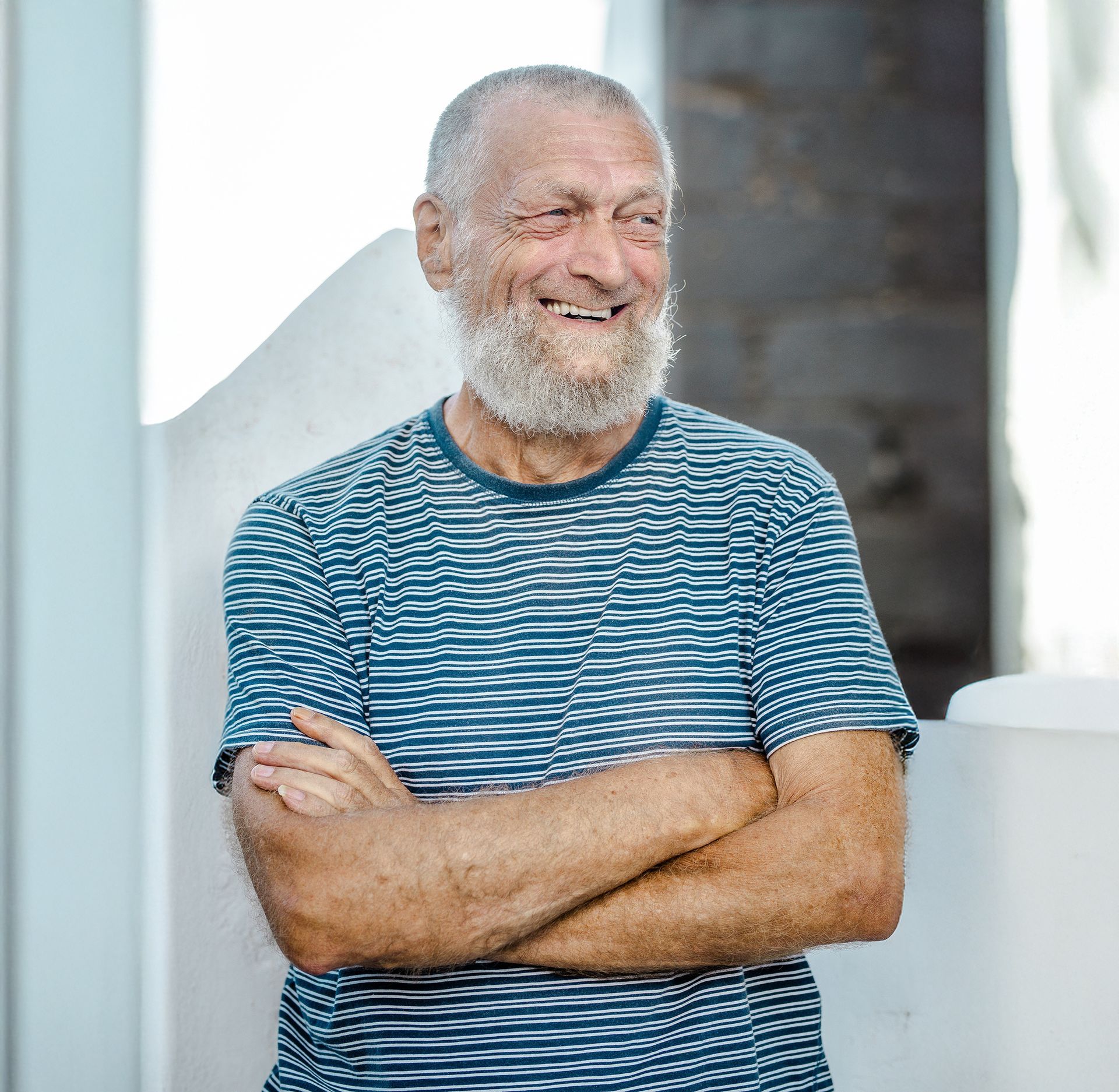 Man with a white beard smiles, arms crossed, wearing a blue and white striped shirt outdoors.