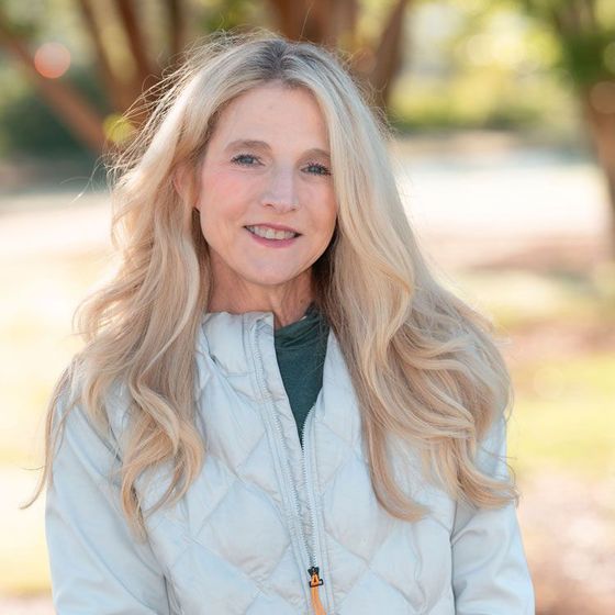 A woman with long blonde hair is wearing a white jacket and smiling for the camera.