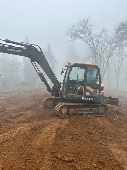 Excavator on muddy construction site in foggy conditions, with bare trees in the background.