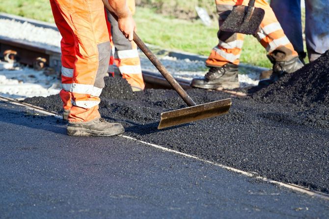 Construction workers in orange high-visibility clothing use tools to spread and level fresh asphalt on a road surface.