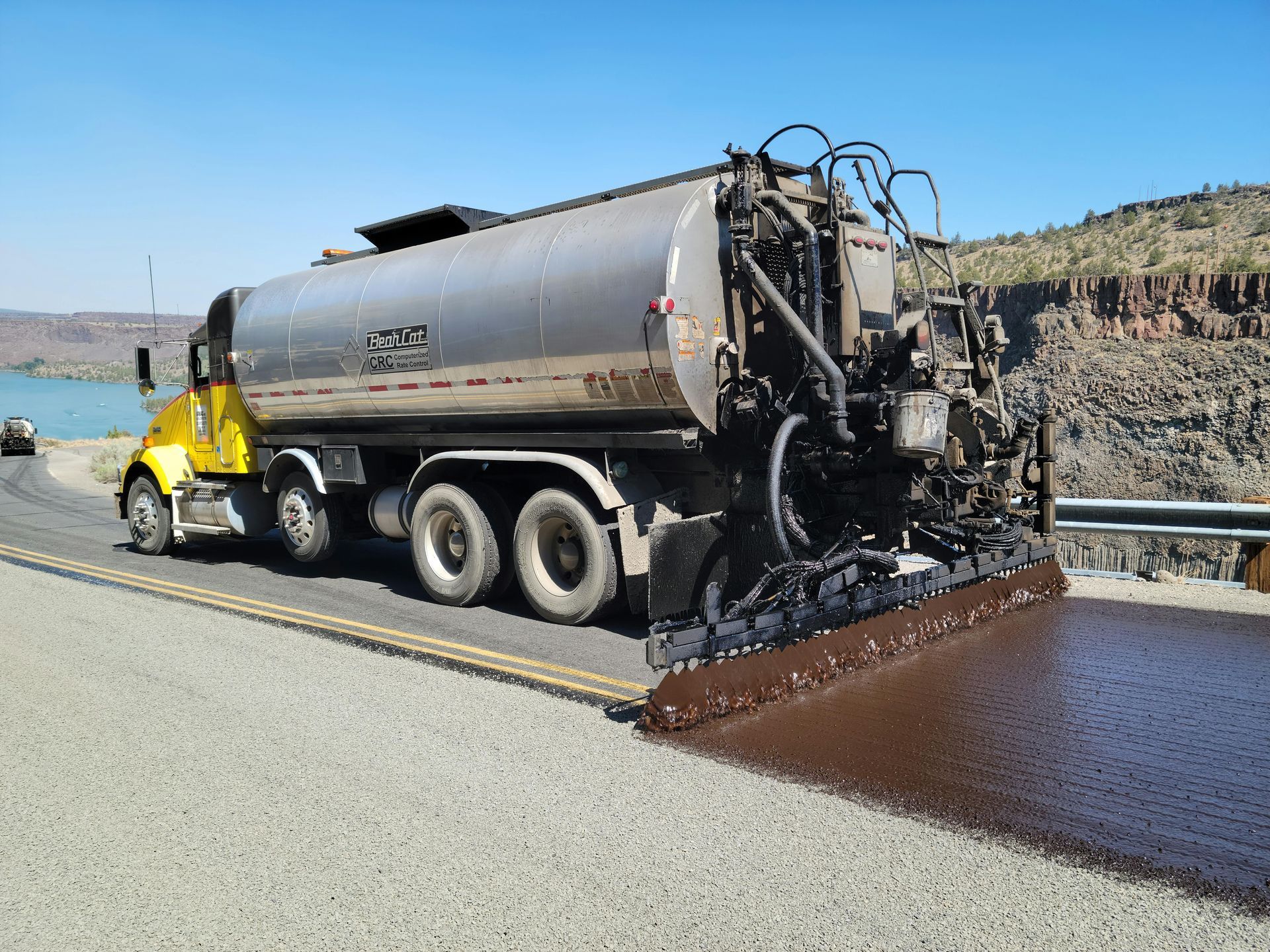 A yellow asphalt distributor truck applies a layer of liquid bitumen to a road surface along a rocky landscape.