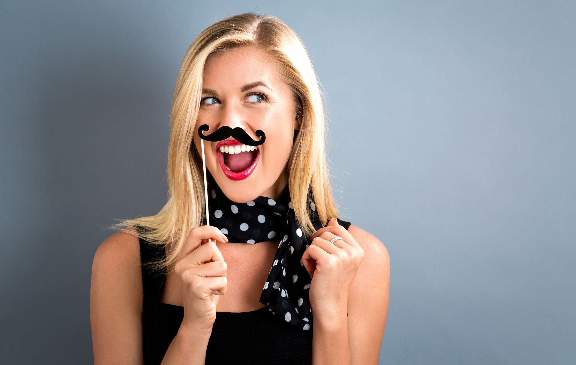 Woman holding a mustache prop with a surprised expression, wearing a black top and polka dot scarf.